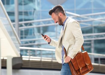 Side portrait of smiling mature man with bag walking outdoors and reading text message on his cell phone