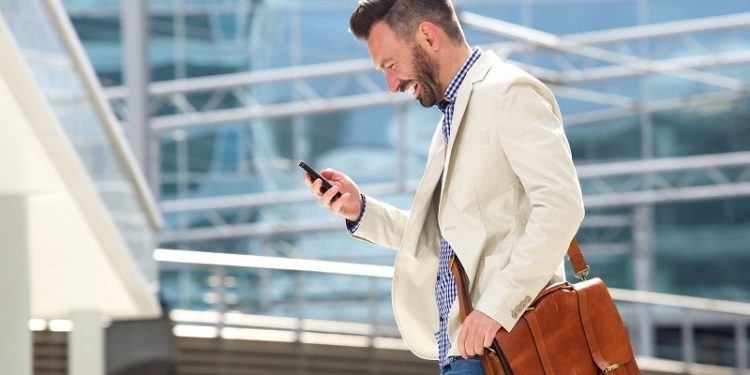 Side portrait of smiling mature man with bag walking outdoors and reading text message on his cell phone