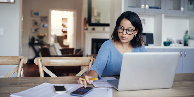 Shot of a young woman using a laptop and calculator while working from home